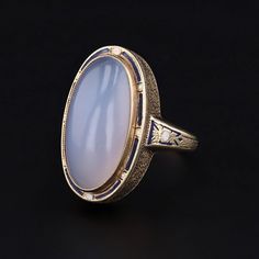 A dramatic blue-hued chalcedony ring accented with blue and white enamel. This gorgeous ring features an ethereal domed chalcedony cabochon measuring 20mm from north to south by 10mm from east to west. The chalcedony is mounted in 14k gold and the stone is framed with alternating blue and white enamel with matching blue and white enamel accents on the sides of the band. The mounting dates to the early 1900s (circa 1910-1920), while the chalcedony is a later replacement stone. The size 6 ring is Elegant Blue Oval Cabochon Moonstone Ring, Oval Cabochon Enamel Ring Collectible, Blue Art Deco Rings With Cabochon, Blue Cabochon Art Deco Rings, Formal Blue Oval Cabochon Moonstone Ring, Blue Cabochon Rings Art Deco Style, Art Deco Blue Cabochon Rings, Formal Blue Chalcedony Ring, Vintage Oval Chalcedony Jewelry