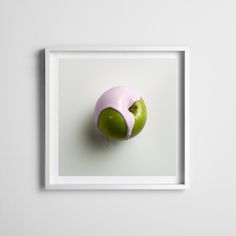 an apple with pink icing on it sitting in front of a white wall mounted frame