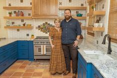 a man and woman standing in the middle of a kitchen with blue cabinets, tile flooring and wooden shelves