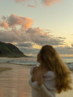 a woman standing on top of a sandy beach next to the ocean at sunset with clouds in the sky