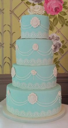 a three tiered blue cake with pink flowers on top and white lace around the edges