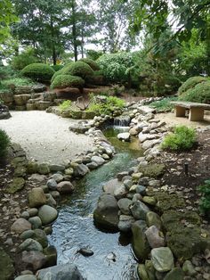 a small stream running through a lush green forest filled with trees and rocks, next to a bench