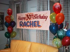 a happy birthday banner with balloons and streamers hanging from the ceiling in front of a couch