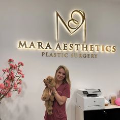 a woman holding a teddy bear in front of a sign that says marra aesthetics plastic surgery