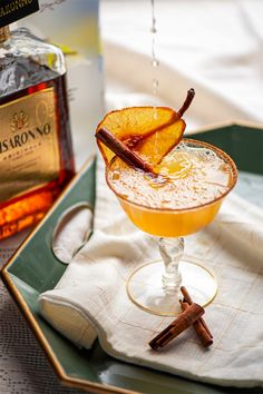 a drink in a glass on a tray with cinnamon and an orange peel garnish