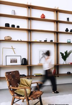 a woman walking past a wooden chair in front of a bookshelf filled with shelves