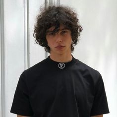 a young man with curly hair wearing a black t - shirt and standing in front of a white wall