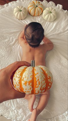 a baby laying on top of a bed next to two pumpkins and a person's hand