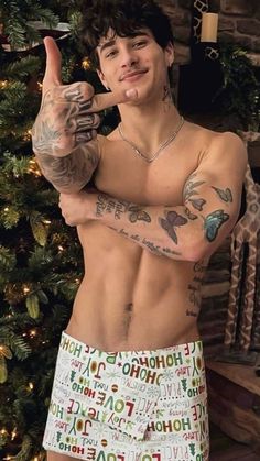 a shirtless man with tattoos giving the peace sign while standing in front of a christmas tree