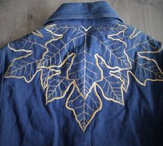 the back of a denim jacket with embroidered leaves and gold thread on it, sitting on a wooden floor