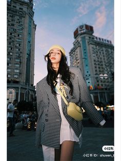 a woman standing in the middle of a city with tall buildings behind her wearing a hat and jacket