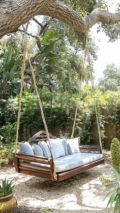 a wooden swing bed hanging from a tree in a backyard with blue pillows on it