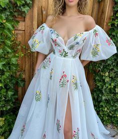 a woman standing in front of a wooden fence wearing a white dress with flowers on it