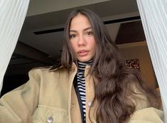 a woman with long dark hair wearing a beige jacket and striped turtle neck sweater, standing in front of a white curtain