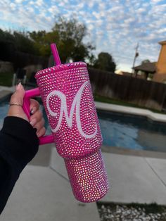 a person holding up a pink tumbler cup with the letter m on it in front of a swimming pool