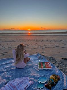 a woman sitting on top of a blue blanket next to the ocean at sunset with donuts