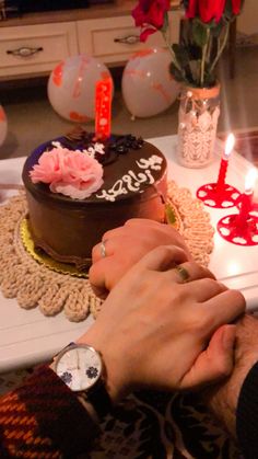 two people holding hands in front of a chocolate cake with roses on it and lit candles