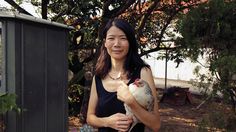 a woman holding a chicken in her right hand while standing next to a fence and trees