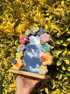 a hand holding up a mirror with flowers on it in front of bushes and blue sky