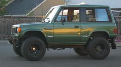 a green four door suv parked in front of a fenced area with a dog sitting in the window