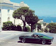 an old car driving down the road in front of a large white building with trees