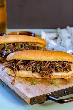 two barbecue sandwiches sitting on top of a cutting board next to a glass of beer