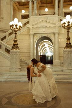 two women in white dresses are hugging each other at the bottom of stairs and chandeliers