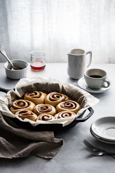 a pan filled with cinnamon rolls on top of a table next to cups and saucers