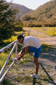 a man bending over to pick up his shoes on the side of a road near a fence