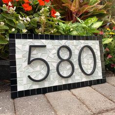 a house number sign sitting on top of a brick sidewalk next to flowers and plants