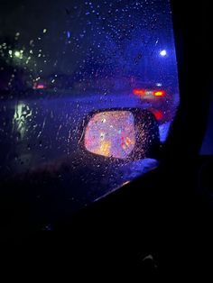 rain is falling on the windshield of a car at night with street lights in the background