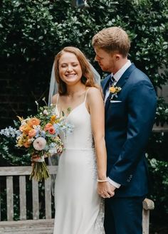 a bride and groom standing next to each other in front of a bench with greenery