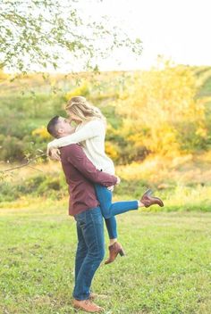 a man holding a woman in his arms while standing on the grass with trees behind him