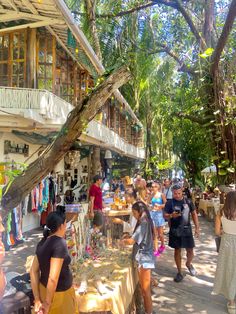 many people are shopping at an outdoor market in the sun, with trees and buildings behind them