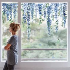 a woman standing in front of a window with blue flowers hanging from it