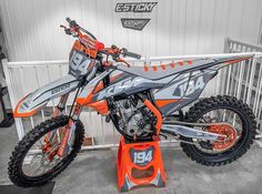 an orange and gray dirt bike parked in front of a white building with the number 34 on it