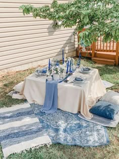 a table set up with blue candles and place settings on a rug in front of a house