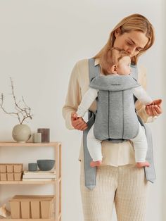 a woman is holding a baby in a grey and white carrier while standing next to a shelf