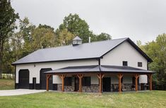 a large white barn with black roof in the middle of a grassy field next to trees