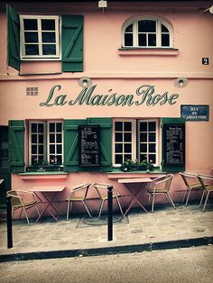 a pink building with green shutters on the windows and tables in front of it