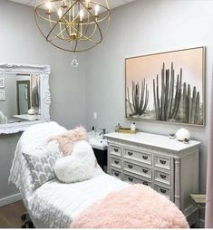 a white bed sitting under a chandelier next to a mirror and dresser in a bedroom