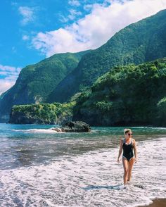 a woman in a bathing suit walking into the water on a beach with mountains in the background