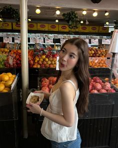 a woman standing in front of a fruit stand holding a bowl of food and looking at the camera