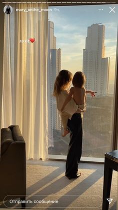 a woman holding a baby standing in front of a window with the city skyline behind her