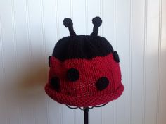 a red and black knitted lady bug hat on top of a wooden stand next to a white wall