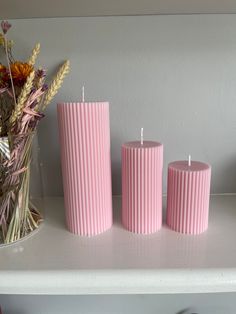 three pink candles sitting on top of a white shelf next to a vase filled with flowers