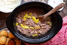 a crock pot filled with beef and peppers next to potatoes on a wooden table