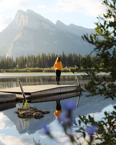 a woman in an orange jacket is standing on a dock near the water and mountains