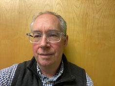 a man wearing glasses and a vest standing in front of a wood paneled wall