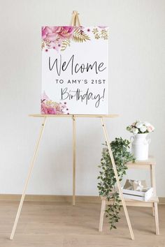 a welcome sign with pink flowers on it next to a wooden easel and potted plant